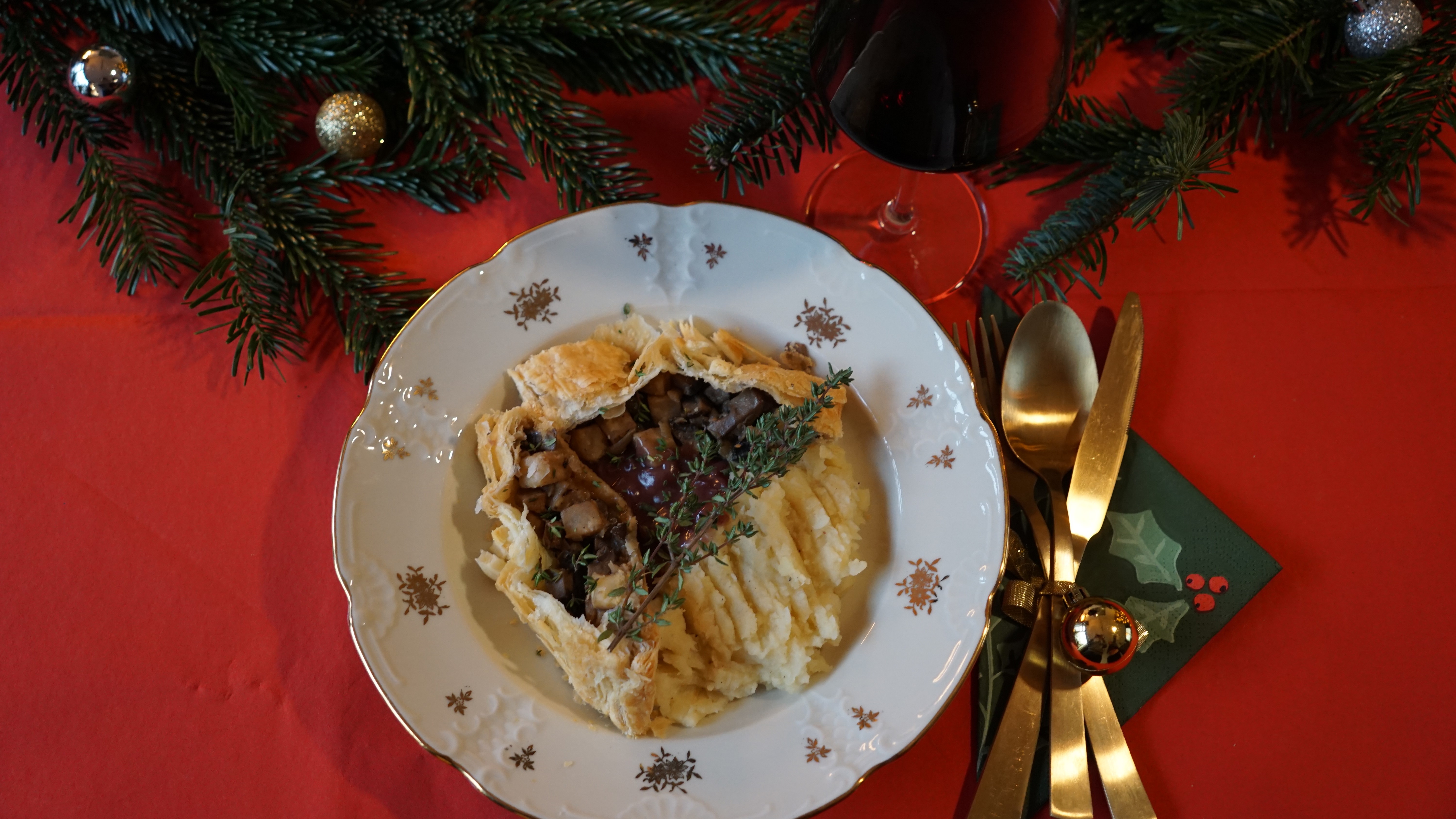 Bladerdeegrol met kastanjechampginons en knolselderij met daarnaast pastinaakpuree en rode wijnsaus op een bord met gouden versiering op een gedekte tafel in kerstsfeer